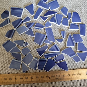 May include: A collection of irregular, smooth-edged blue ceramic shards with white undersides, scattered above a wooden ruler that measures up to 150 millimetres, showing various shapes and sizes of these broken pottery pieces for mosaic projects.