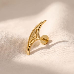 May include: A gold-toned metal body jewellery piece featuring a curved, crescent-like shape with intricate cut-out detailing along the inner edge. The piece includes a flat circular backing post, designed for insertion into a piercing, with a polished finish.