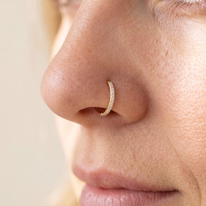 May include: A gold-toned nose hoop ring featuring a row of small, clear, sparkling stones embedded along the outer edge. The circular jewellery is positioned securely through the nostril, showcasing a polished metallic finish and a delicate design.