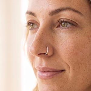 May include: A silver-toned nose ring hoop featuring a row of small, clear, sparkling stones embedded along the outer edge. The circular jewelry piece is positioned securely in the nostril, showcasing a polished metallic finish and clear gems.