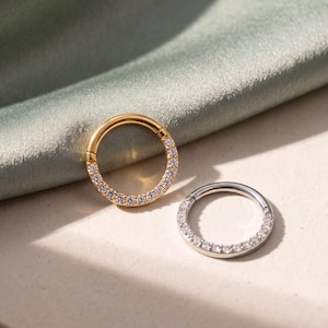 May include: Two circular hoop rings, one in a gold tone and one in a silver tone, each featuring a row of small, clear, sparkling stones embedded along the front half of the circular band, creating a continuous shimmering effect on the metal.
