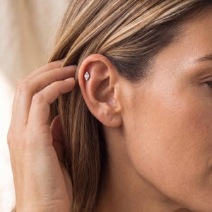 May include: A small, silver-toned metal stud earring in a diamond shape, featuring a central clear stone. The earring is positioned in the upper cartilage area of the ear, secured with a post backing, showcasing a clean and simple geometric design.