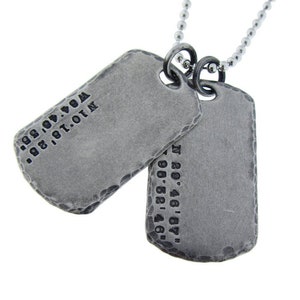 以下が含まれることがあります： 銀色のチェーンネックレスに、銀色のドッグタグが2つ付いています。ドッグタグには、座標が刻印されています。上のタグには「N 18.181.01N W 0.01.01.00M」と刻印されています。下のタグには「N 80.46.67.00 W 00.68.41.46」と刻印されています。