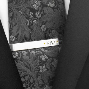 Op de afbeelding: Een zilveren stropdas met de initialen "KAA" in gouden letters. De stropdas is op een zwarte stropdas met patroon, met een zwart colbert in de achtergrond. De stropdas heeft een bloemenpatroon.