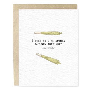 May include: A birthday card with a white background and a brown envelope. The card features two illustrations of rolled joints and the text "I USED TO LIKE JOINTS BUT NOW THEY HURT happy birthday."
