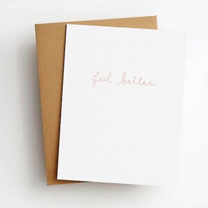 May include: A white greeting card with the words "feel better" in a delicate, cursive font. The card is layered over a brown kraft paper envelope, creating a simple, elegant design. The background is a clean, white surface.