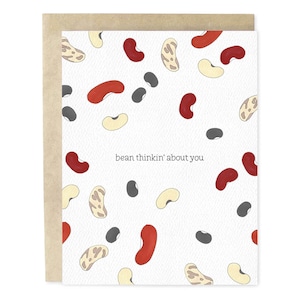 May include: A greeting card with a white background featuring a pattern of illustrated beans in red, beige, black, and yellow. The text "bean thinkin' about you" is printed in black on the card. The card is inside a brown envelope.