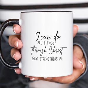 Peut inclure: Une tasse à café en céramique blanche avec une poignée noire. La tasse porte une inscription noire qui dit "Je peux tout faire par le Christ qui me fortifie."