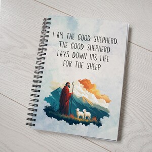 Könnte beinhalten: Ein spiralgebundenes Notizbuch mit einem Aquarellhintergrund, auf dem ein Hirte mit einem Stab und zwei Schafe zu sehen sind. Der Text auf dem Cover lautet "Ich bin der gute Hirte. Der gute Hirte gibt sein Leben für die Schafe."