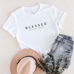 Può includere: Maglietta bianca con la scritta "Blessed to be God's daughter" stampata in nero.