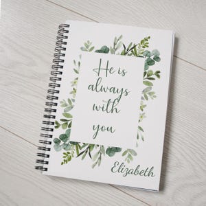 Op de afbeelding: Een wit spiraalnotitieblok met een groen bloemmotief en de tekst "He is always with you" in een zwart kader. De naam "Elizabeth" staat onderaan het notitieblok.
