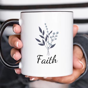 Peut inclure: Une tasse en céramique blanche avec une anse et un bord noirs. La tasse présente un motif floral dans les tons bleus et gris avec le mot "Foi" imprimé en noir sous le motif.