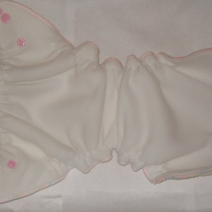 Puede incluir: Pa&ntilde;al de tela blanco con ribete rosa y broches. El pa&ntilde;al est&aacute; doblado por la mitad y tiene un borde rizado.