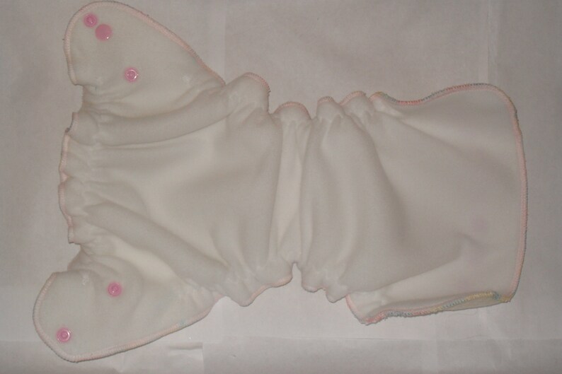 Puede incluir: Pa&ntilde;al de tela blanco con ribete rosa y broches. El pa&ntilde;al est&aacute; doblado por la mitad y tiene un borde rizado.