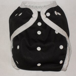 May include: A black cloth diaper with white trim and white snaps. The diaper has a white trim around the edges and white snaps along the front.