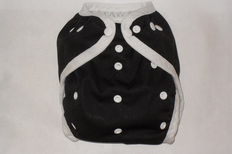 May include: A black cloth diaper with white trim and white snaps. The diaper has a white trim around the edges and white snaps along the front.