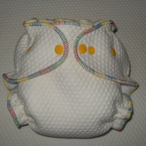 Può includere: Un pannolino di stoffa bianco con un bordo colorato e bottoni gialli. Il pannolino è fatto di un tessuto morbido e testurizzato.
