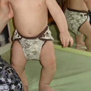 May include: A baby wearing a brown and white patterned cloth diaper with a brown elastic waistband.