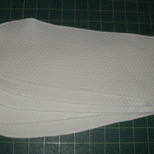 Puede incluir: Cinco insertos de pañales de tela reutilizables de algodón blanco apilados. Los insertos están hechos de una tela suave y texturizada.