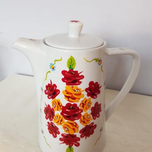 May include: White ceramic teapot with a lid and handle, decorated with a vibrant floral pattern. The flowers are mainly red and yellow, with green leaves and small blue and yellow accents. The teapot has a spout for pouring.