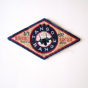 May include: A diamond-shaped patch with a blue border and red stitching. The patch features a white background with a blue circle containing the text "TANGO RANGE" and a silhouette of a jet plane. The text "3/3 AUTHENTIQUE TRAINING" is on the left side of the circle and "LINE FLIGHTGEAR MOSCOW" is on the right side.