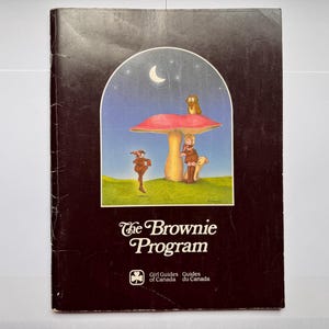 May include: A black book titled The Brownie Program with a central illustration featuring a large red mushroom, a crescent moon, an owl, and two figures in a grassy field. The cover includes the Girl Guides of Canada logo and text.