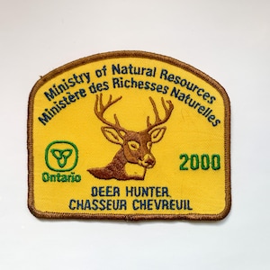Könnte beinhalten: Ein gelber Aufnäher mit braunem Rand, der einen Hirschkopf und Geweih zeigt. Der Text lautet "Ministry of Natural Resources", "Ontario", "2000" und "Deer Hunter / Chasseur Chevreuil". Mit einem grün-weißen Logo.