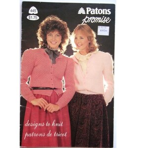 Peut inclure: Une couverture de livre de modèles de tricot vintage représentant deux femmes portant des cardigans roses. Le livre est intitulé "Patons Promise" et porte une étiquette de prix de 1,75 $. La couverture comprend également le texte "designs to knit patrons de tricot".
