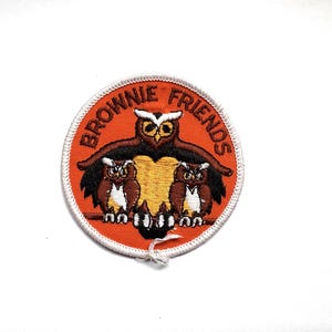 Peut inclure: Écusson rond avec un fond orange et les mots "BROWNIE FRIENDS" en lettres orange. L'écusson représente trois hiboux bruns, blancs et jaunes. L'écusson a une bordure cousue blanche.