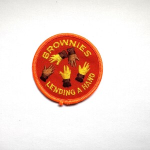 Può includere: Una toppa rotonda arancione delle Brownies con le parole "BROWNIES LENDING A HAND" ricamate in un motivo curvo. La toppa presenta mani ricamate in varie tonalità di pelle, con guanti gialli e marroni.