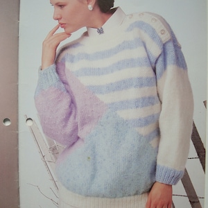 Peut inclure: Un pull ray&eacute; bleu clair, blanc et violet avec un motif g&eacute;om&eacute;trique. Le pull a un col rond et des manches longues.