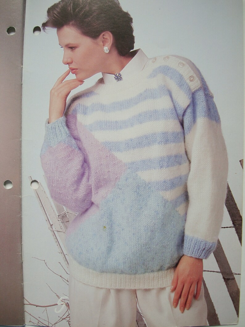 Peut inclure: Un pull ray&eacute; bleu clair, blanc et violet avec un motif g&eacute;om&eacute;trique. Le pull a un col rond et des manches longues.