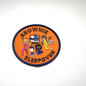 May include: Circular embroidered patch with the text "BROWNIE SLEEPOVER". The design features cartoon girls painting, wearing pyjamas, on an orange background with a navy blue border. Ideal for a Brownie's uniform.