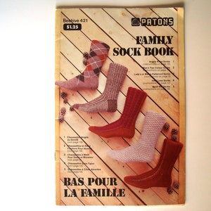 May include: A vintage "Family Sock Book" featuring knitted sock patterns. The cover displays images of different sock styles, including argyle and textured designs. The book is on a wooden background.