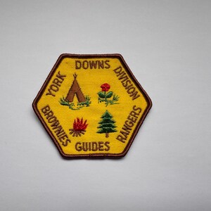 May include: A yellow embroidered patch with a brown border. The patch features a tent, a rose, a campfire, and a pine tree. The text on the patch reads "York Downs Division Brownies Guides Rangers".