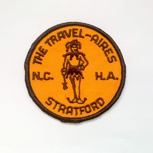 May include: A circular patch with a brown border and an orange centre. The patch features a brown illustration of a jester and the text "THE TRAVEL-AIRES N.C. H.A. STRATFORD" in brown lettering.