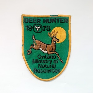 Può includere: Toppa ricamata a forma di scudo verde con bordo dorato. La toppa presenta un cervo che salta, un sole giallo e il testo "DEER HUNTER 1978". Sotto il cervo c'è il testo "Ontario Ministry of Natural Resources".