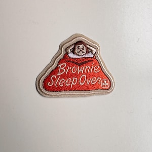 Puede incluir: Parche bordado con una figura de dibujos animados bajo una manta roja. El parche tiene un borde beige y las palabras "Brownie Sleep Over" en escritura blanca. Un pequeño emblema está en la esquina inferior derecha. Para un grupo de Brownies.