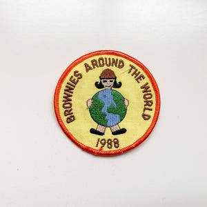 Może przedstawiać: Haftowana naszywka z napisem "BROWNIES AROUND THE WORLD" i rokiem "1988". Naszywka przedstawia postać z kreskówki trzymającą globus. Naszywka ma żółte tło z czerwoną obwódką.
