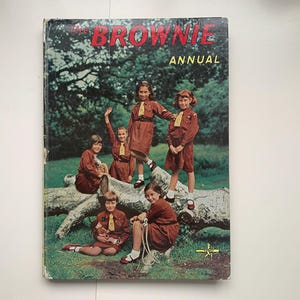 Può includere: Copertina del libro vintage "The Brownie Annual" con un gruppo di ragazze in uniforme marrone con cravatte gialle. Il titolo in lettere rosse su uno sfondo di alberi.