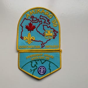 Può includere: Toppa ricamata in due parti con sfondo azzurro e bordi dorati. La sezione superiore presenta una mappa del Canada con una foglia d'acero rossa e un giglio dorato. Sono visibili anche le scritte "AMIKARO 99" e "DORCHESTER ONTARIO".
