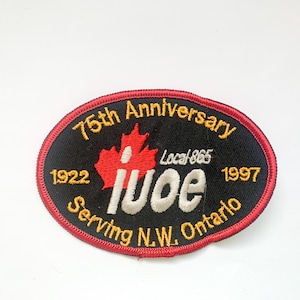 Może przedstawiać: Okrągła naszywka z czarnym tłem i czerwoną obwódką. Na naszywce widnieje napis "75th Anniversary" i "Serving N.W. Ontario". Widoczny jest również czerwony liść klonu i napis "IUOE". Zawiera lata 1922 i 1997.
