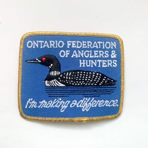 May include: Embroidered patch with a blue background and a gold border. The patch features a loon illustration and text that reads "ONTARIO FEDERATION OF ANGLERS & HUNTERS" and "I'm making a difference."