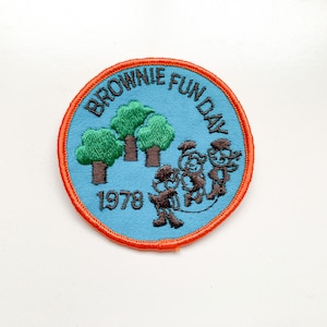 Può includere: Toppa ricamata con la scritta "BROWNIE FUN DAY 1978". La toppa presenta uno sfondo blu con alberi verdi e figure marroni che saltano la corda. La toppa ha un bordo arancione.
