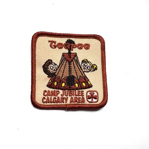 Könnte beinhalten: Gestickter Aufnäher mit braunem Rand und beigem Hintergrund. Der Aufnäher zeigt ein Tipi-Design mit zwei Cartoon-Gesichtern auf jeder Seite. Der Text "Teepee" steht oben und "CAMP JUBILEE CALGARY AREA" unten.