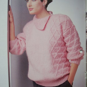 Peut inclure: Un pull rose en tricot avec un motif de tricot torsad&eacute; sur les manches et un col. Le pull est port&eacute; par une personne aux cheveux courts et fonc&eacute;s.