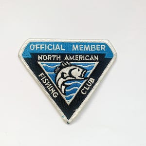 May include: A diamond-shaped patch with the words "OFFICIAL MEMBER NORTH AMERICAN FISHING CLUB" in white and blue. A white fish is jumping out of the water in the center of the patch. The patch has a blue border.