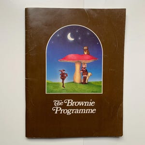 Peut inclure: Un livret marron intitulé "The Brownie Programme" avec une illustration colorée. L'illustration présente une scène fantaisiste avec un champignon, une fille, un chien, un hibou et une figurine dansant sous un ciel nocturne avec un croissant de lune.