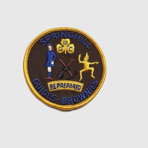 May include: A circular Springhill Guides-Brownies patch with a gold border and dark brown centre. The patch features a girl in a blue dress, a yellow figure, and the words "BE PREPARED".