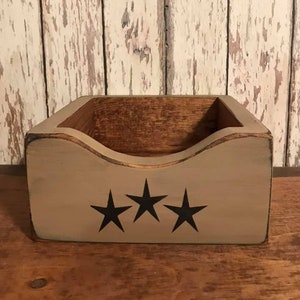 May include: A wooden box with a curved top edge and three black star designs. The box is painted in a neutral color with a distressed finish, and sits on a wooden surface. The background is a distressed white wooden wall.